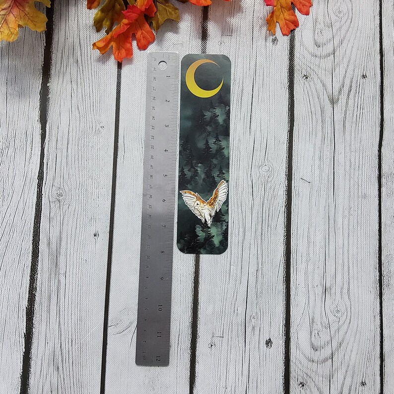 May include: A dark green bookmark with a white owl flying in front of a crescent moon and a dark forest. The bookmark is 6 inches tall.