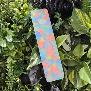 May include: A rectangular bookmark with a colorful, abstract design of overlapping, semi-circular shapes in shades of blue, pink, yellow, and orange. The bookmark is set against a backdrop of lush green foliage.
