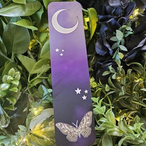 May include: A bookmark with a celestial design featuring a crescent moon, stars, and a butterfly. The background transitions from purple to black, creating a night sky effect. The butterfly has detailed wing patterns.