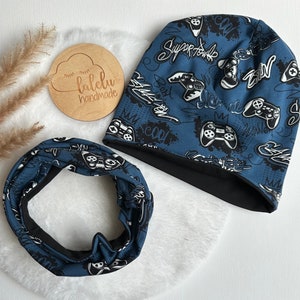 Könnte beinhalten: Ein blaues und schwarzes, umkehrbares Mützen- und Halstuch-Set mit einem Gamer-Motiv. Der Druck zeigt weißen Text und Bilder von Videospielcontrollern. Der Text enthält "Super Fowls", "Best", "Awesome", "My Cool", "Play", "Winner" und "Level Up".