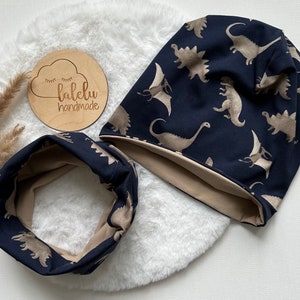 Könnte beinhalten: Ein Set aus zwei dunkelblauen und beigen Strickaccessoires mit Dinosaurier-Motiv. Das Set beinhaltet eine Mütze und einen Halsschmuck. Die Mütze hat einen umgeschlagenen Saum. Der Halsschmuck ist ein Schlaufenmodell. Die Accessoires sind handgefertigt von "lalelu handmade".