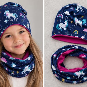 May include: A navy blue beanie and matching infinity scarf set featuring a repeating pattern of white unicorns, colorful rainbows, pink clouds, and yellow stars. Both items are lined with a solid bright pink fabric on the interior.