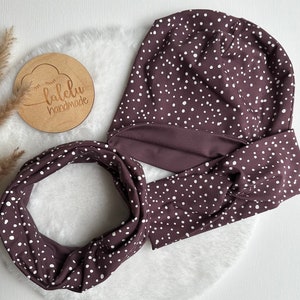 Könnte beinhalten: Eine braune und weiße Polka-Dot-Mütze und ein passender Infinity-Schal. Die Mütze hat einen umgeschlagenen Rand und der Schal ist aus einem weichen, dehnbaren Stoff.
