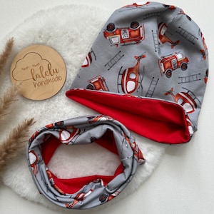 May include: A gray and red reversible beanie and neck warmer set with a fire truck and helicopter print. The set features a red interior and a gray exterior with a white background. The print includes red fire trucks, helicopters, and ladders.