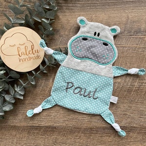 Könnte beinhalten: Ein graues und türkisfarbenes Baby-Kuscheltuch in Form eines Nilpferds mit dem Namen "Paul" auf der Vorderseite gestickt. Das Kuscheltuch hat ein Sternmuster und weiße Akzente. Ein Holzschild mit dem Text "lalelu handmade" befindet sich im Hintergrund.
