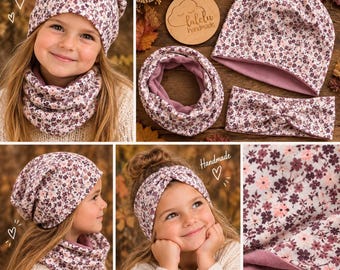 May include: A set consisting of a beanie, a neck warmer, and a headband, all featuring a floral pattern with small pink and dark purple flowers on a light background. The items are lined with a solid mauve fabric, and a wooden tag reads lalelu handmade.