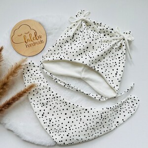 May include: White baby hat and bib with black polka dots. The hat has ties and the bib is a bandana style. The hat and bib are handmade and the label says "lalelu handmade".