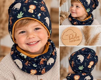 May include: A dark blue beanie and matching infinity scarf set featuring a repeating pattern of cartoon elephants, lions, and green foliage. The accessories have a solid mustard yellow lining and are displayed with a wooden logo tag reading lalelu handmade.