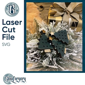 May include: Three dark green wooden Christmas tree ornaments with gold stars and the words "Grecian" and "Eliza" on them. The ornaments are sitting on a bed of white flocked greenery.