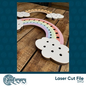 May include: Two rainbow-coloured wooden yarn holders with cloud-shaped bases. The rainbow holders have holes for yarn and are layered in shades of pink, orange, and yellow. The clouds are white and have holes for yarn.