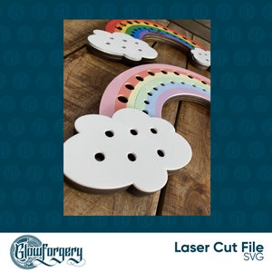 May include: A rainbow-coloured wooden rainbow with a white cloud attached to the bottom. The rainbow has holes for threading string or yarn. The cloud also has holes for threading string or yarn. The rainbow is made of multiple layers of wood, each painted a different colour of the rainbow. The cloud is white and has a smooth surface.