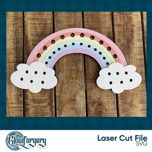May include: A wooden rainbow cutout with clouds on either side. The rainbow has holes for threading string or yarn. The clouds also have holes for threading. The rainbow is pink, orange, yellow, green, blue, and purple. The clouds are white. The background is a wooden surface.