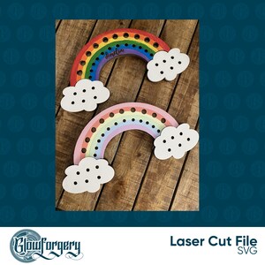 May include: Two rainbow-coloured wooden rainbow shapes with clouds. The larger rainbow is painted in red, orange, yellow, green, blue, indigo, and violet. The smaller rainbow is painted in pink, light blue, lavender, and yellow. Both rainbows have holes for threading string or yarn. The larger rainbow has the name "Amelia" written on it.
