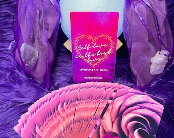 May include: A pink and purple affirmation deck with a rose design and a pink gem. The deck is titled "Self-love is the best love" and is by Brandi Taylor.