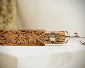 May include: A brown leather keychain with a floral design and a silver metal clasp. The keychain has a decorative silver metal button with a floral design.