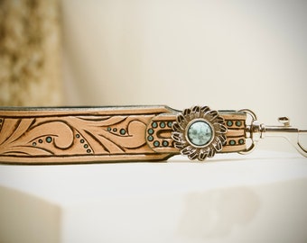May include: A light brown leather key chain with a floral design and a silver flower charm with a turquoise stone in the center. The key chain has a silver swivel hook.