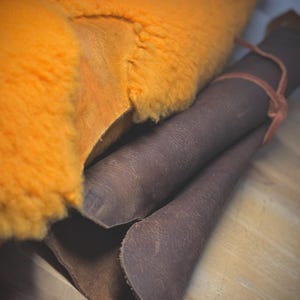 May include: An image showing orange sheepskin and rolled brown leather. The sheepskin has a fluffy texture. The leather is rolled up and tied with a thin, reddish-brown band. The materials are on a light-coloured wooden surface.