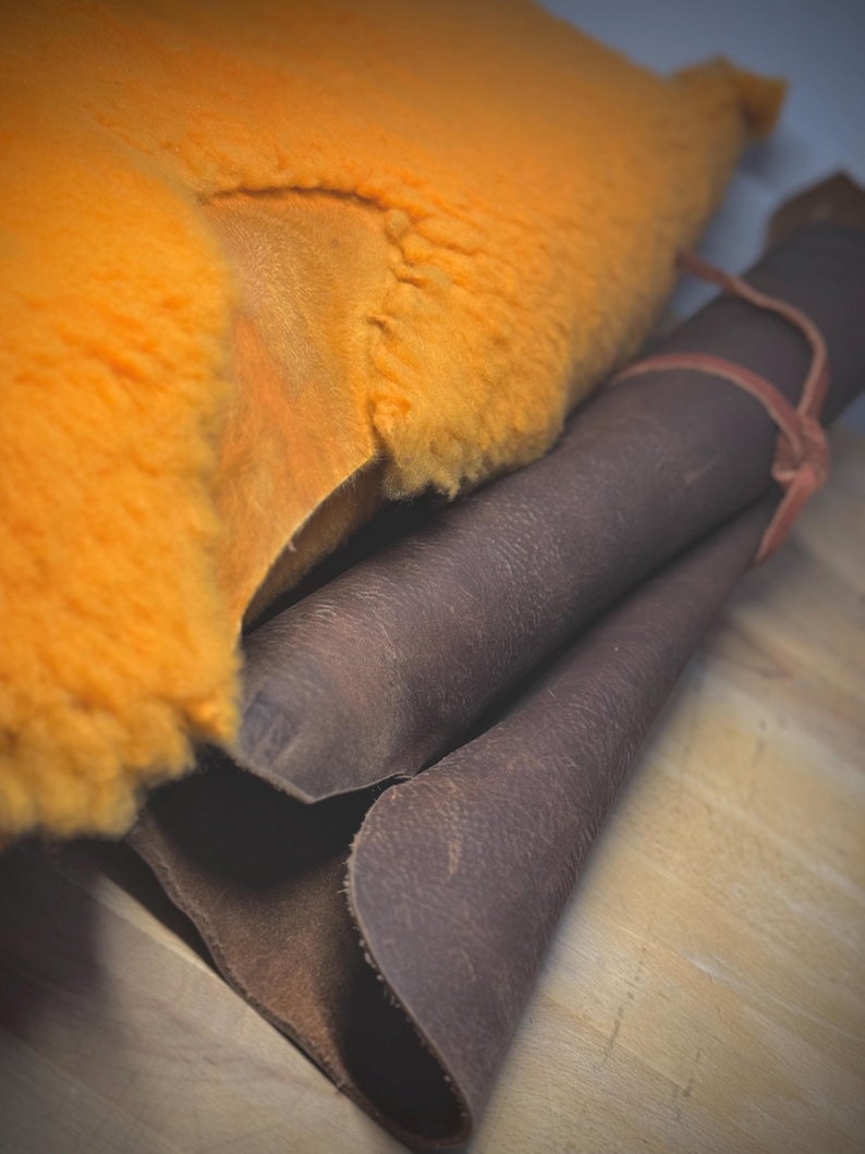 May include: An image showing orange sheepskin and rolled brown leather. The sheepskin has a fluffy texture. The leather is rolled up and tied with a thin, reddish-brown band. The materials are on a light-coloured wooden surface.