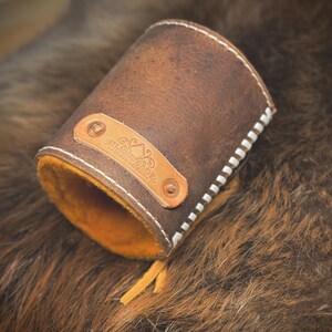 May include: A brown leather cuff with white stitching and an orange interior. A small leather tag with text and rivets is attached. The cuff is open at one end and has a small leather tassel.