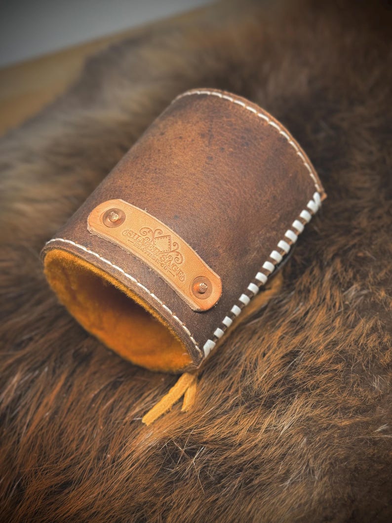 May include: A brown leather cuff with white stitching and an orange interior. A small leather tag with text and rivets is attached. The cuff is open at one end and has a small leather tassel.