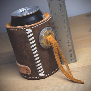 May include: A brown leather can cooler with white stitching and a decorative silver and gold emblem. The cooler has a leather strap and holds a black can. A ruler is next to the cooler, measuring approximately 7.6 cm.