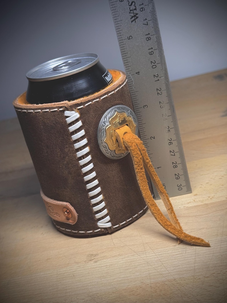 May include: A brown leather can cooler with white stitching and a decorative silver and gold emblem. The cooler has a leather strap and holds a black can. A ruler is next to the cooler, measuring approximately 7.6 cm.