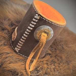 May include: A brown leather cylinder with white stitching and an orange interior. A decorative silver and gold emblem is attached to the side, with a tan leather strap hanging down. The item is resting on a brown, furry surface.