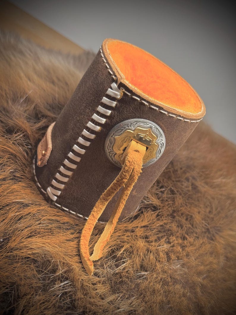 May include: A brown leather cylinder with white stitching and an orange interior. A decorative silver and gold emblem is attached to the side, with a tan leather strap hanging down. The item is resting on a brown, furry surface.