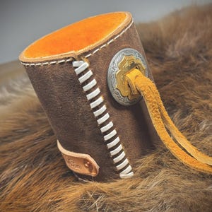 May include: A brown leather cup holder with white stitching and an orange interior. It features a decorative silver and gold emblem and a yellow leather tassel. The cup holder is designed to hold a beverage.