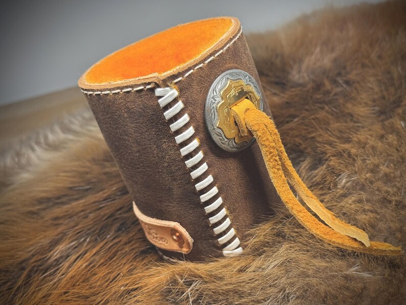 May include: A brown leather cup holder with white stitching and an orange interior. It features a decorative silver and gold emblem and a yellow leather tassel. The cup holder is designed to hold a beverage.
