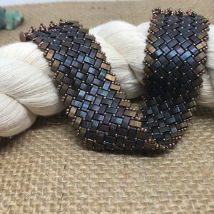 May include: A woven bracelet made of dark blue and gold beads. The bracelet is on a white and brown background.