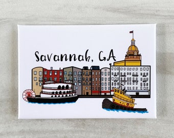 May include: A white magnet with a colorful illustration of the Savannah, GA skyline. The illustration includes buildings, a riverboat, and a tugboat. The text "Savannah, GA" is written in black cursive.