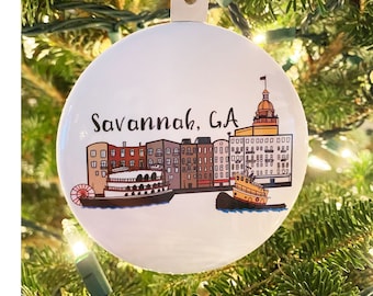 Puede incluir: Un adorno navideño redondo blanco con una ilustración colorida del horizonte de Savannah, Georgia. El adorno presenta edificios, un río y dos barcos. El texto "Savannah, GA" está impreso en el adorno.