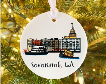 Puede incluir: Adorno navideño redondo de cerámica blanca con una colorida ilustración del horizonte de Savannah, GA, que incluye un barco de vapor y un remolcador. El texto "Savannah, GA" está escrito en escritura negra. El adorno está suspendido por una cinta blanca.