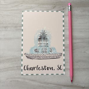 May include: A postcard featuring a blue and black illustration of a fountain with a pineapple on top, with the text "Charleston, SC". A pink pencil rests beside the postcard.