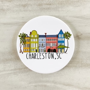 May include: A white round coaster with a colorful illustration of a row of houses in Charleston, South Carolina. The text "CHARLESTON, SC" is printed below the houses.