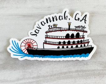 May include: A white sticker with a black and white illustration of a steamboat with a red paddle wheel. The text "Savannah, GA" is written in black cursive above the boat. The text "Nancy's Designs" is written in black below the boat.