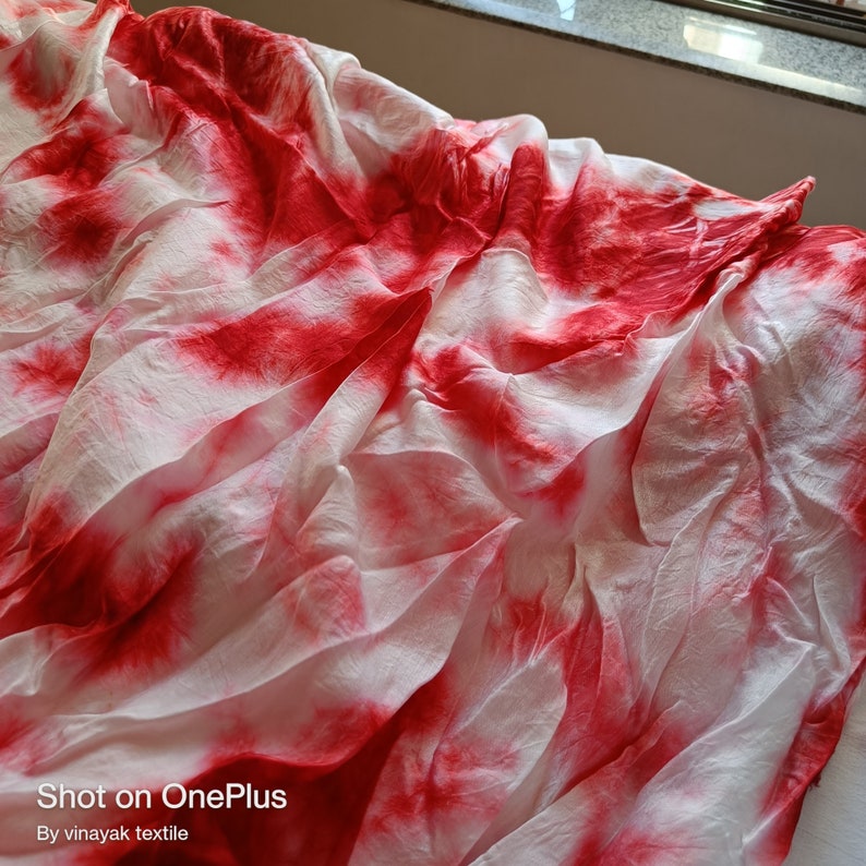 May include: White fabric with red tie-dye pattern. The fabric is crumpled and has a soft, flowing texture. "Shot on OnePlus" and "By vinayak textile" are printed at the bottom of the image.