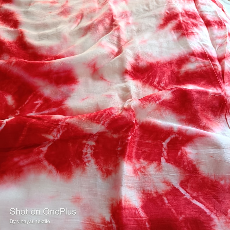 May include: White fabric with red tie-dye pattern. The fabric is wrinkled and has a soft, flowing texture. "Shot on OnePlus" and "By vinayak textile" are printed at the bottom of the image.