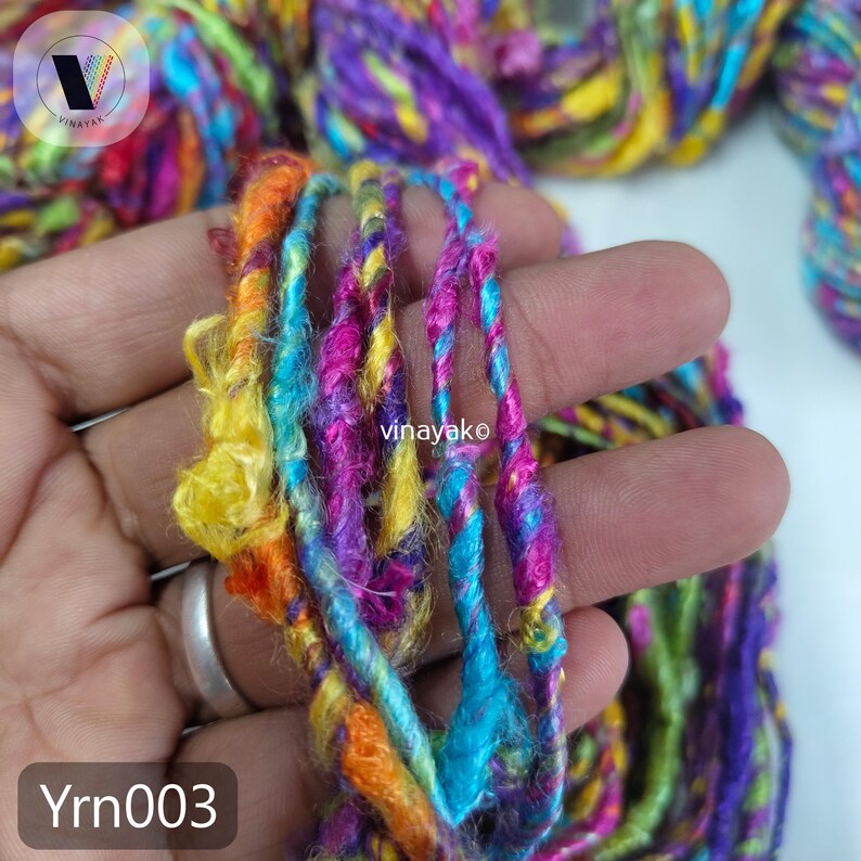 May include: Close-up of colorful yarn strands in shades of blue, purple, yellow, and orange. The yarn is held in a hand, with the text "vinayako" and "Yrn003" visible. The yarn appears to be a blend of textures.
