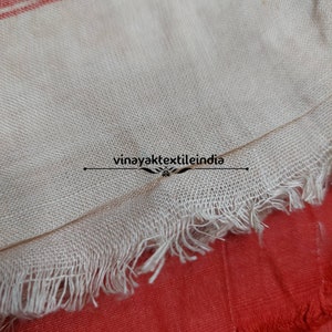 Pode incluir: Close-up de um tecido bege com uma borda com franjas, sobre um fundo vermelho. O tecido tem uma textura tecida e uma borda crua e inacabada. O texto "vinayaktextileindia" &eacute; vis&iacute;vel.