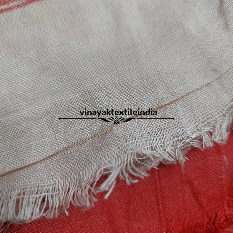 Pode incluir: Close-up de um tecido bege com uma borda com franjas, sobre um fundo vermelho. O tecido tem uma textura tecida e uma borda crua e inacabada. O texto "vinayaktextileindia" &eacute; vis&iacute;vel.