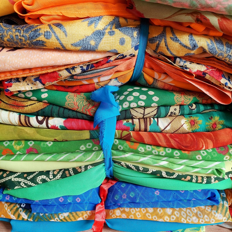 May include: A stack of folded fabric pieces in various colours including orange, yellow, green, and blue, featuring diverse floral and geometric patterns. The layers are secured together by a vertical blue fabric tie and a red fabric tie at the bottom.