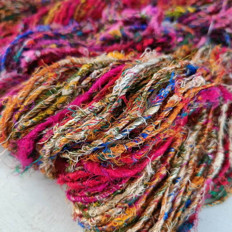 K&ouml;nnte beinhalten: Nahaufnahme eines bunten Garns aus recycelter Sari-Seide. Das Garn ist eine Mischung aus Rosa, Orange, Gelb, Gr&uuml;n, Blau und Wei&szlig;. Das Garn ist weich und texturiert und eignet sich perfekt zum Stricken oder H&auml;keln.