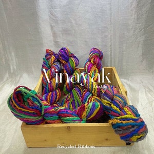 May include: A wooden box filled with colourful recycled ribbon yarn. The yarn is made from upcycled fabric scraps and is available in a variety of colours. The box is labelled "Vinayak Textile" and "Recycled Ribbons".