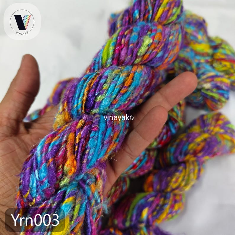 May include: A close-up of a skein of colorful yarn. The yarn is a mix of purple, blue, orange, and green. The text "Yrn003" is visible in the bottom left corner. The yarn is held in a hand.