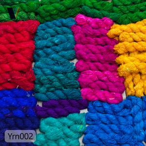 Puede incluir: Una colorida variedad de hilos en varios tonos de verde, rojo, azul, morado, rosa y amarillo. La lana está dispuesta en un patrón de cuadrícula, con el texto "Yrn002" en la esquina inferior izquierda.