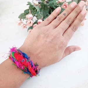 K&ouml;nnte beinhalten: Ein farbenfrohes Armband aus gewebten F&auml;den in den Farben Pink, Rot, Blau, Gr&uuml;n und Gold. Das Armband ist um das Handgelenk einer Person gebunden.