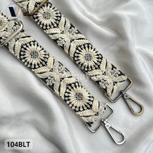 Puede incluir: Dos correas ajustables para bolso con base negra y bordado floral en color crema. Cada correa incluye mosquetones giratorios de metal en tonos plateado y bronce. El texto 104BLT es visible en la esquina inferior izquierda.