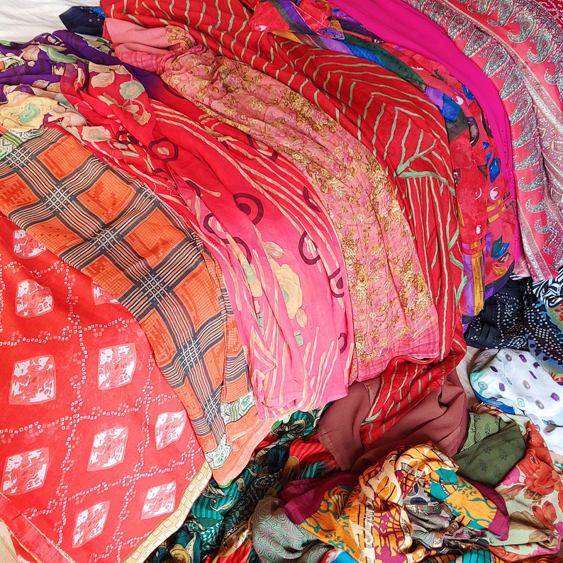 May include: A collection of folded fabric pieces featuring a variety of patterns and colours, including bright red, orange, pink, and purple. The textiles display diverse designs such as floral motifs, geometric plaid, and intricate paisley prints.